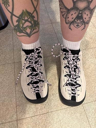 Person wearing unique lace-patterned sneakers, standing on tiled floor with tattooed legs visible