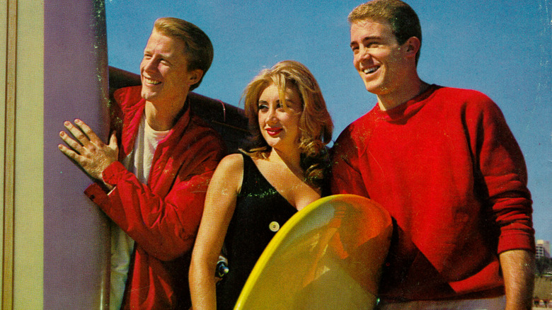 Jan and Dean in red jumpers smiling on beach with surfboards and a young lady