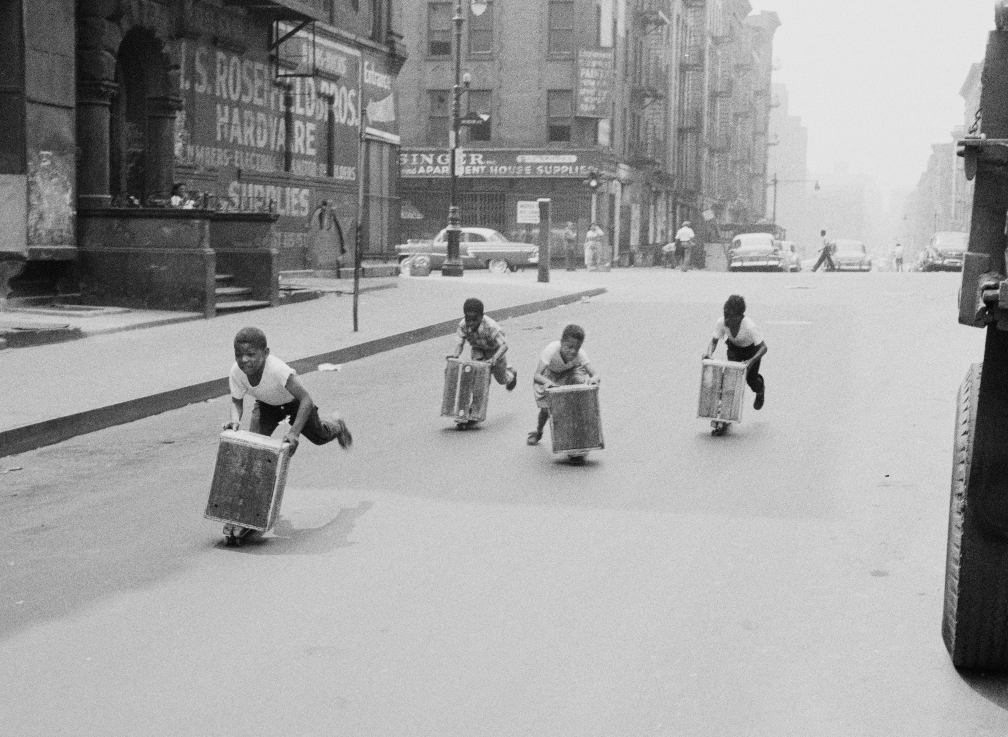 Children ride homemade scooters down an urban street lined with parked cars and buildings, capturing a moment of play in a mid-20th century cityscape