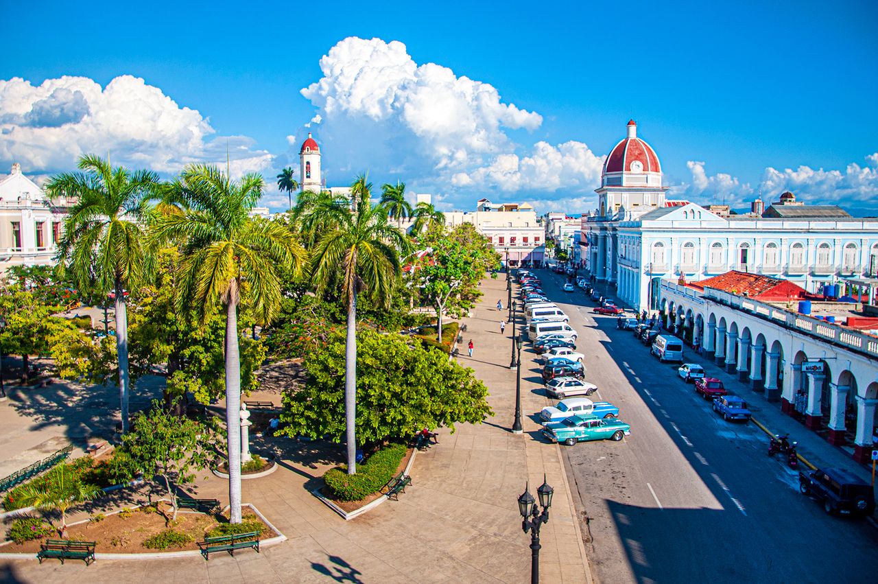 A tropical plaza with palm trees adjacent buildings include a domed structure and parked vintage cars along a street