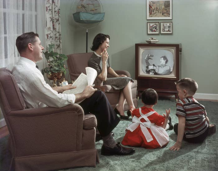 1950s family watching a puppet show on TV; parents seated, children on floor, room with vintage decor