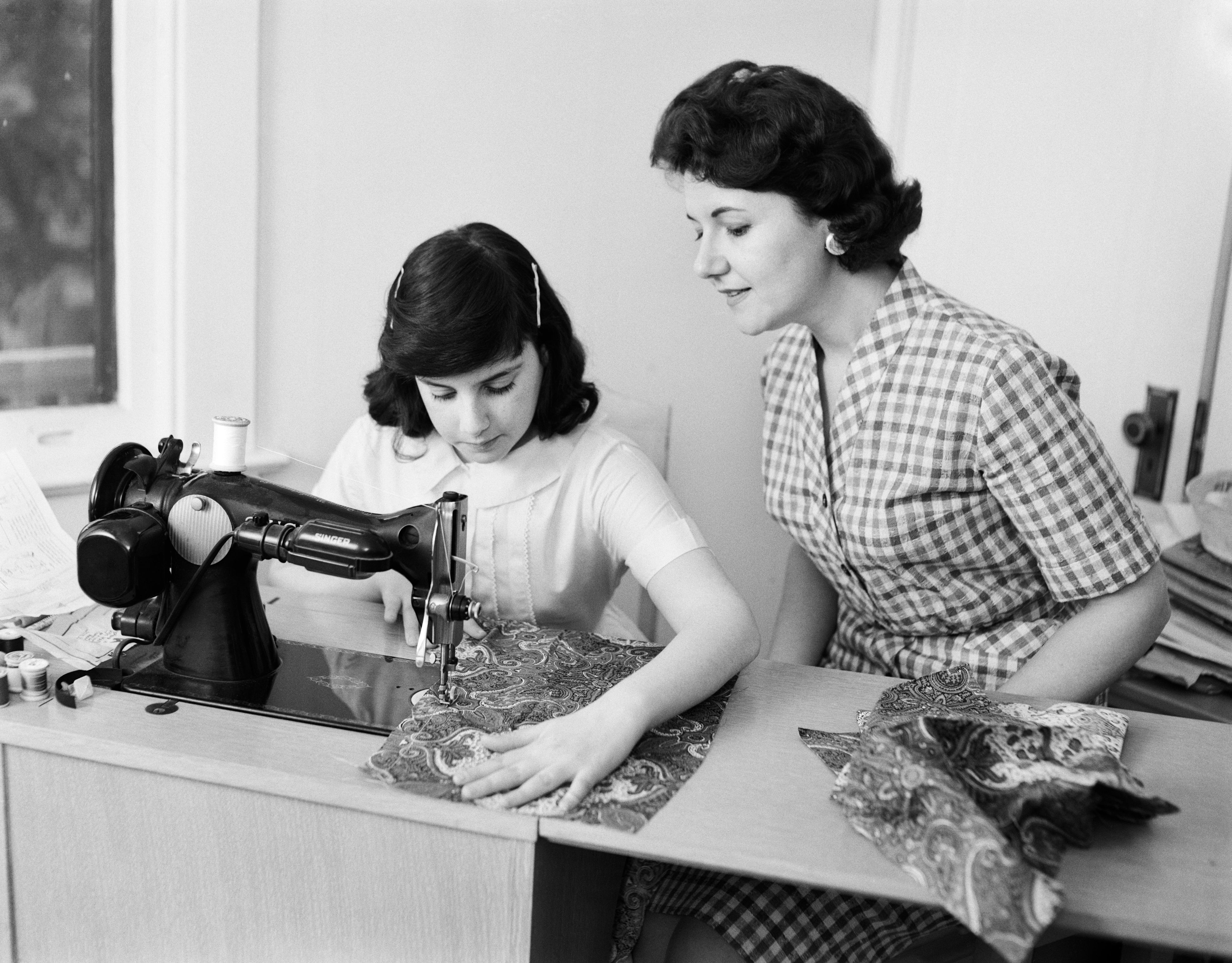 A woman in a checkered dress guides a young girl using a sewing machine, working on a patterned fabric