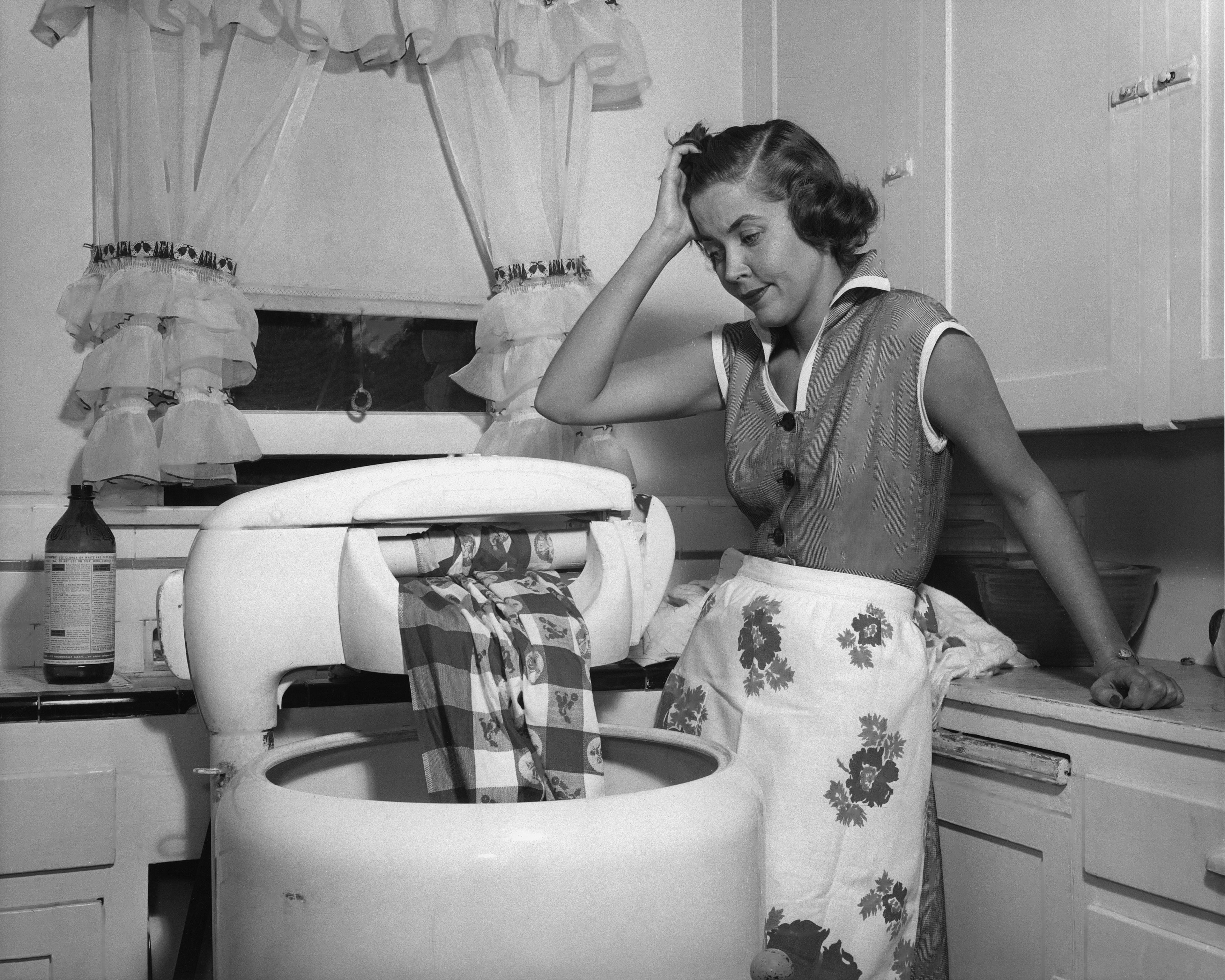 A woman in 1950s attire looks frustrated as she operates a vintage washing machine in a kitchen