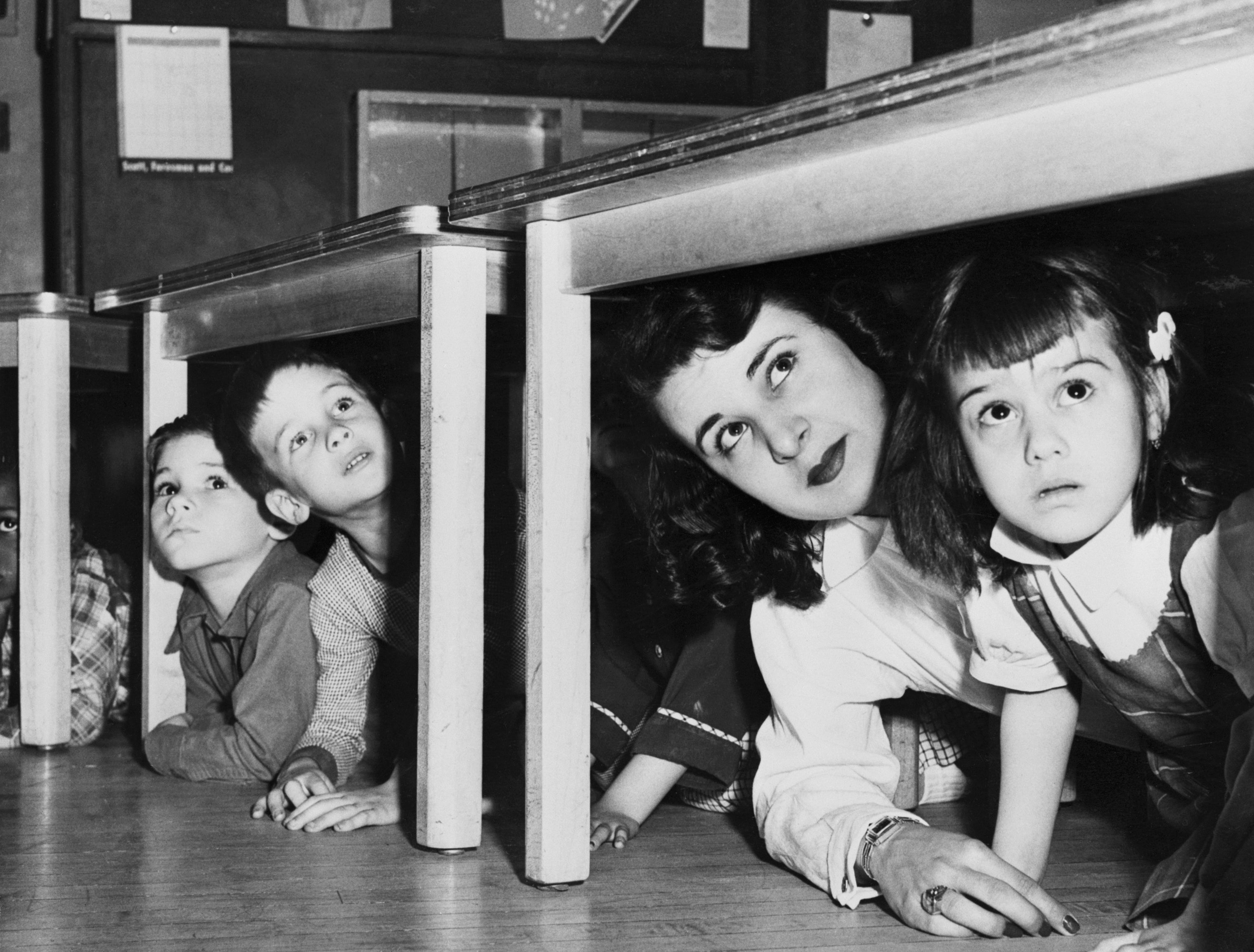Children and a woman practice a duck-and-cover drill under desks, looking ahead attentively