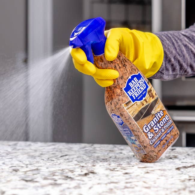 A person wearing yellow gloves sprays Bar Keepers Friend Granite & Stone Cleaner on a countertop