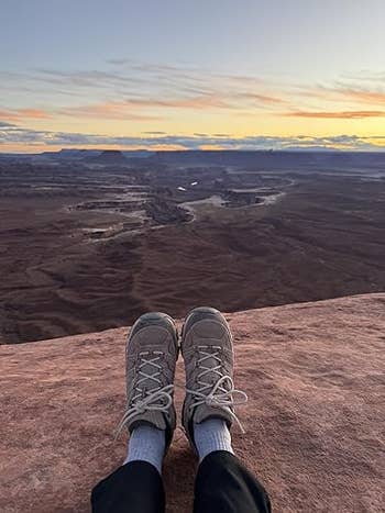Person sitting on a cliff edge with hiking shoes, overlooking a vast canyon landscape at sunset