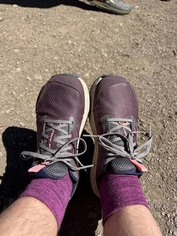 Two feet wearing athletic shoes and purple socks, standing on a gravel surface. Another shoe is visible in the background