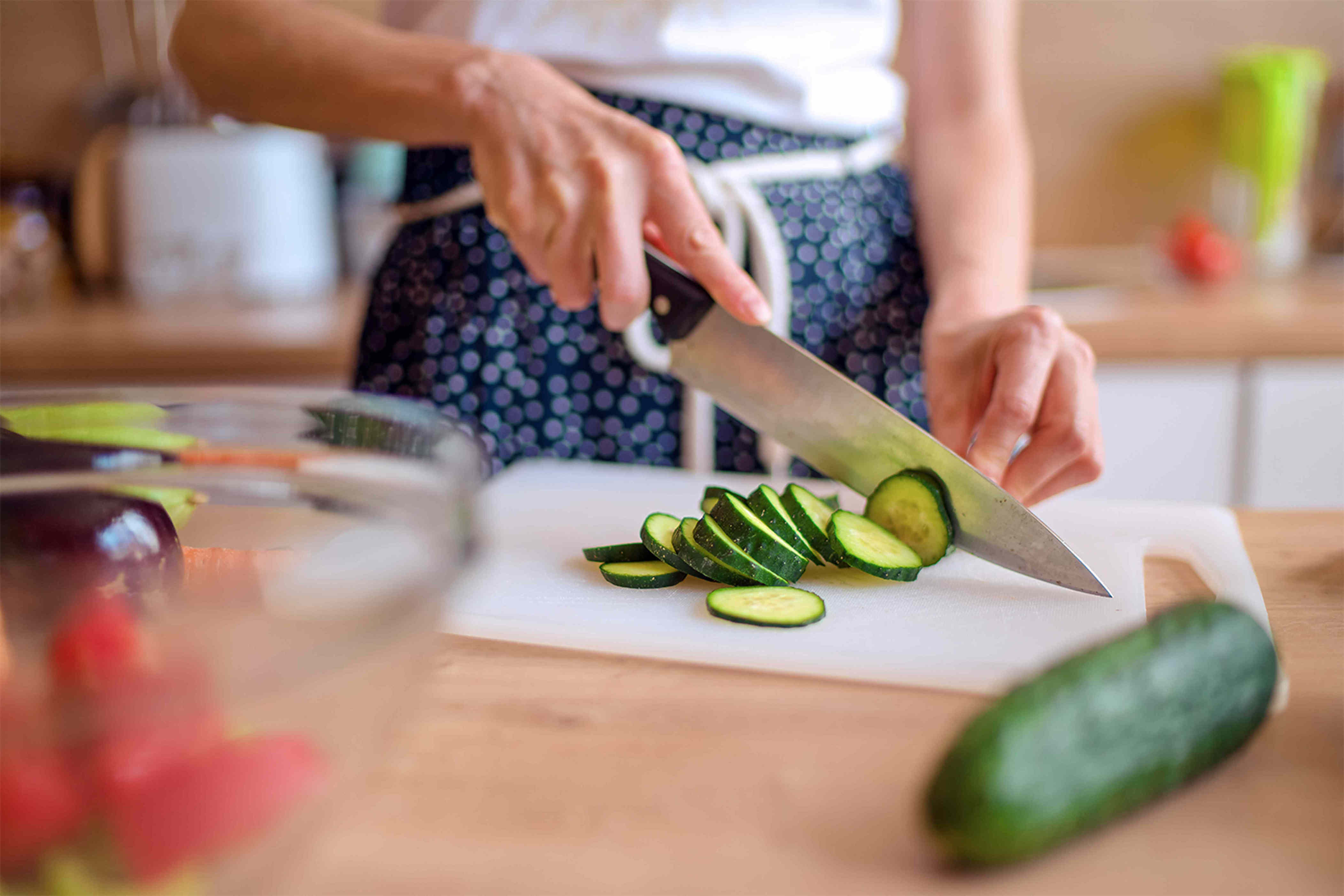 A person slicing a cucumber on a cutting board in a kitchen other vegetables visible nearby