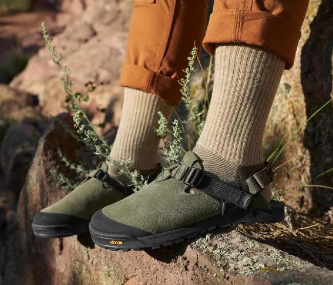 Person wearing beige socks with green sandals and brown pants, standing outdoors on rocks, showcasing practical, rugged footwear style