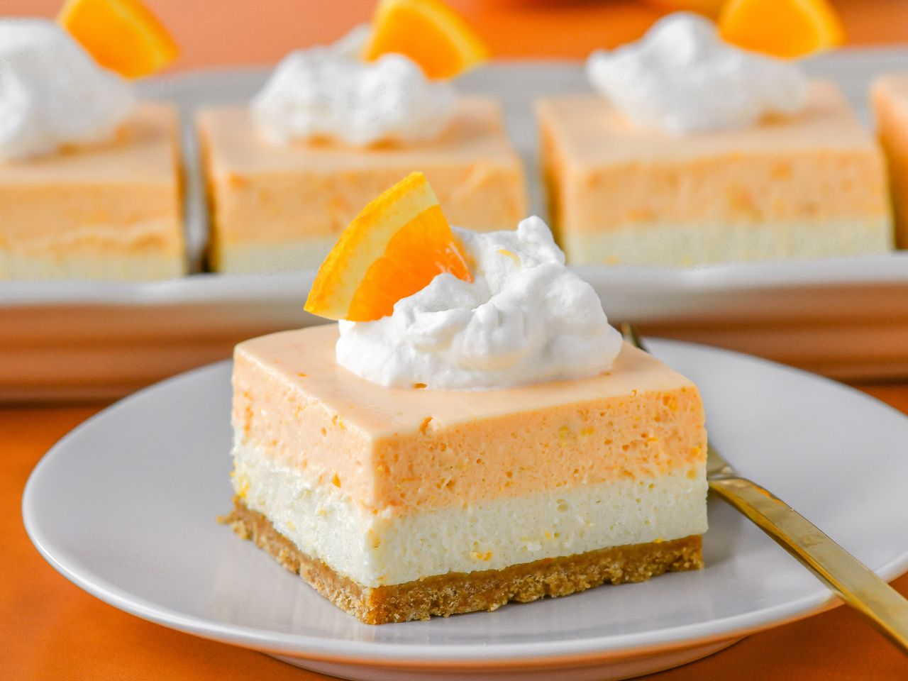 layered creamsicle bar serving on a plate with whipped cream and orange slice; more bars in background