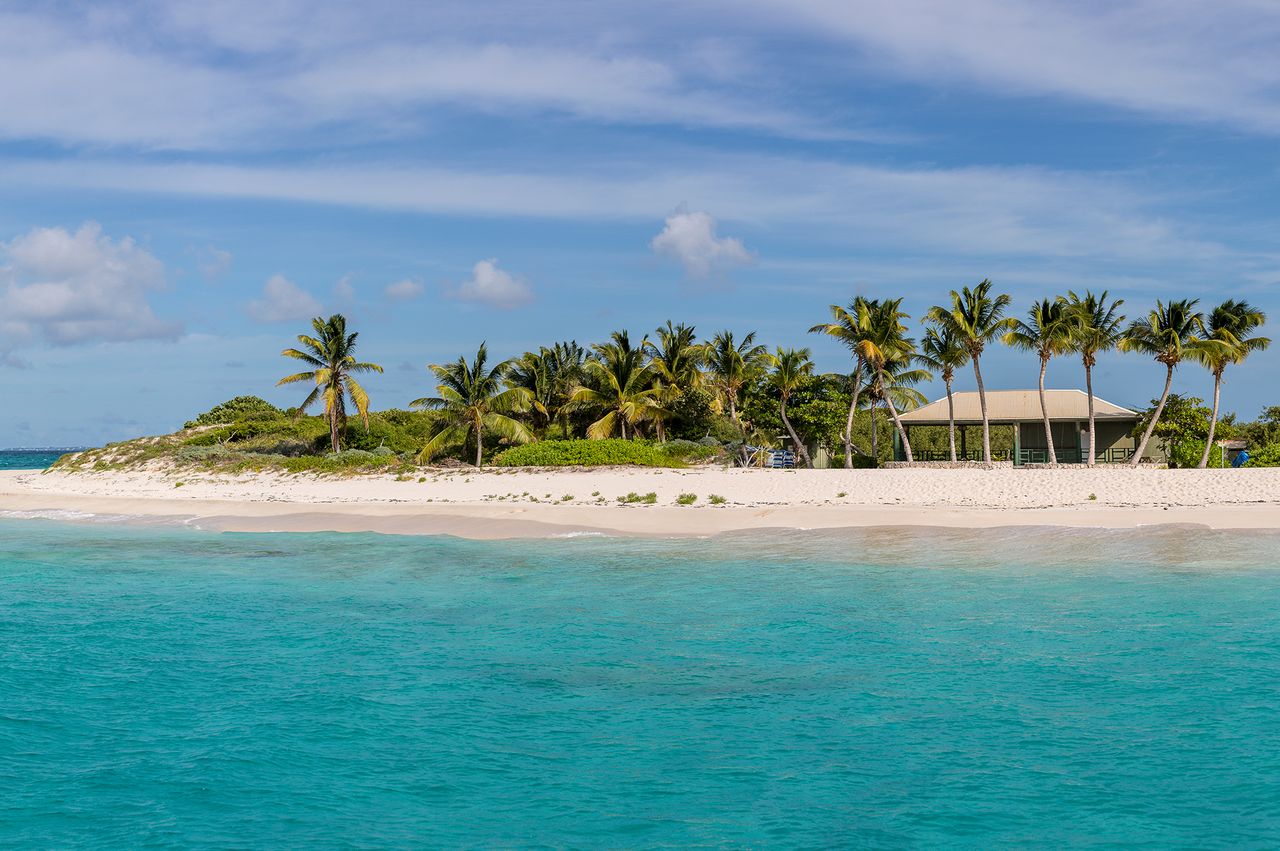 A tropical beach with palm trees and a small building surrounded by turquoise water on a sunny day