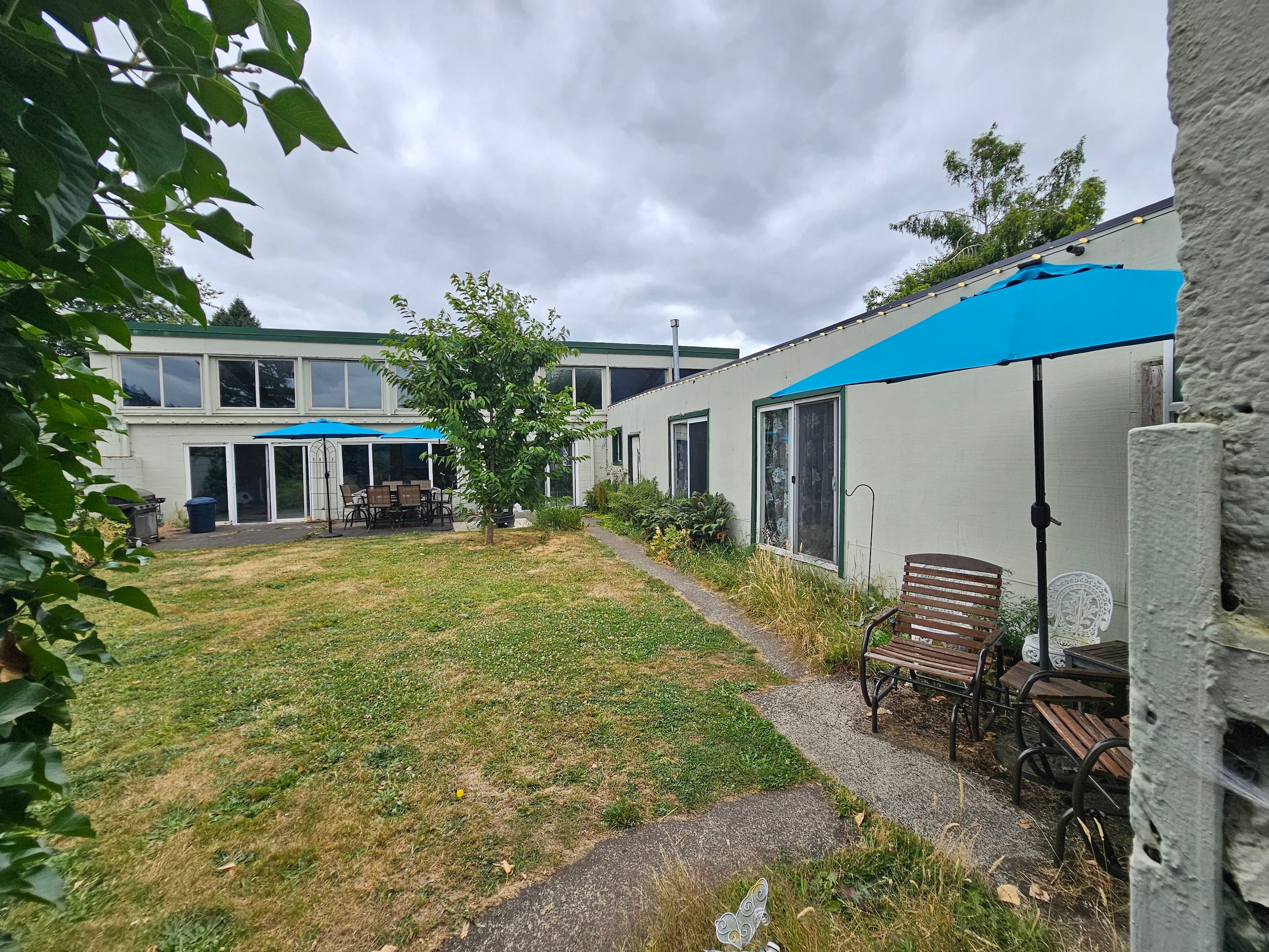 Courtyard outside a light-colored building with sliding doors, blue patio umbrellas, benches, trees, and a cloudy sky.