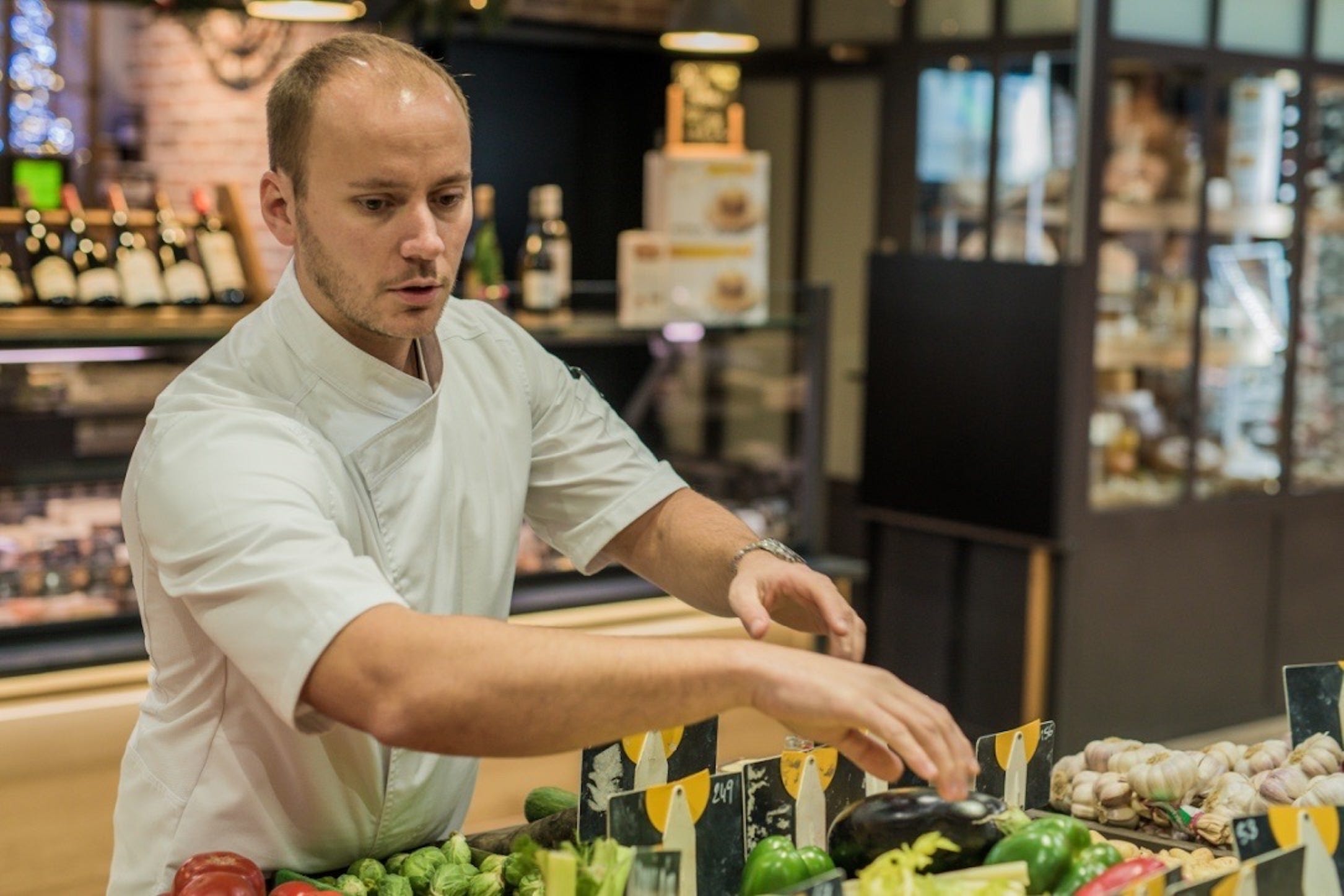 Chef in a white uniform arranges fresh vegetables at a market produce counter.