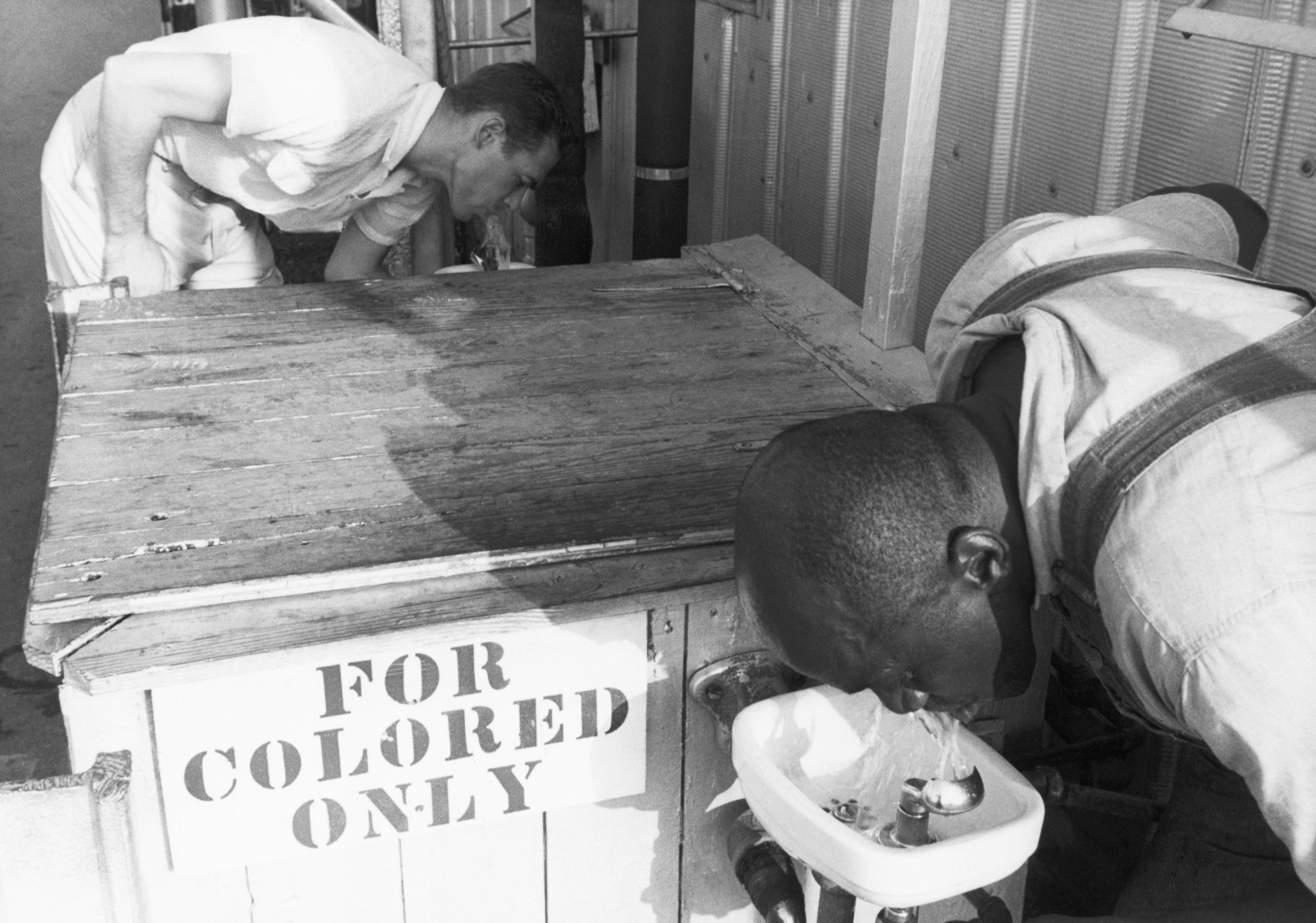 Two men drink from separate water fountains; a "For Colored Only" sign highlights racial segregation