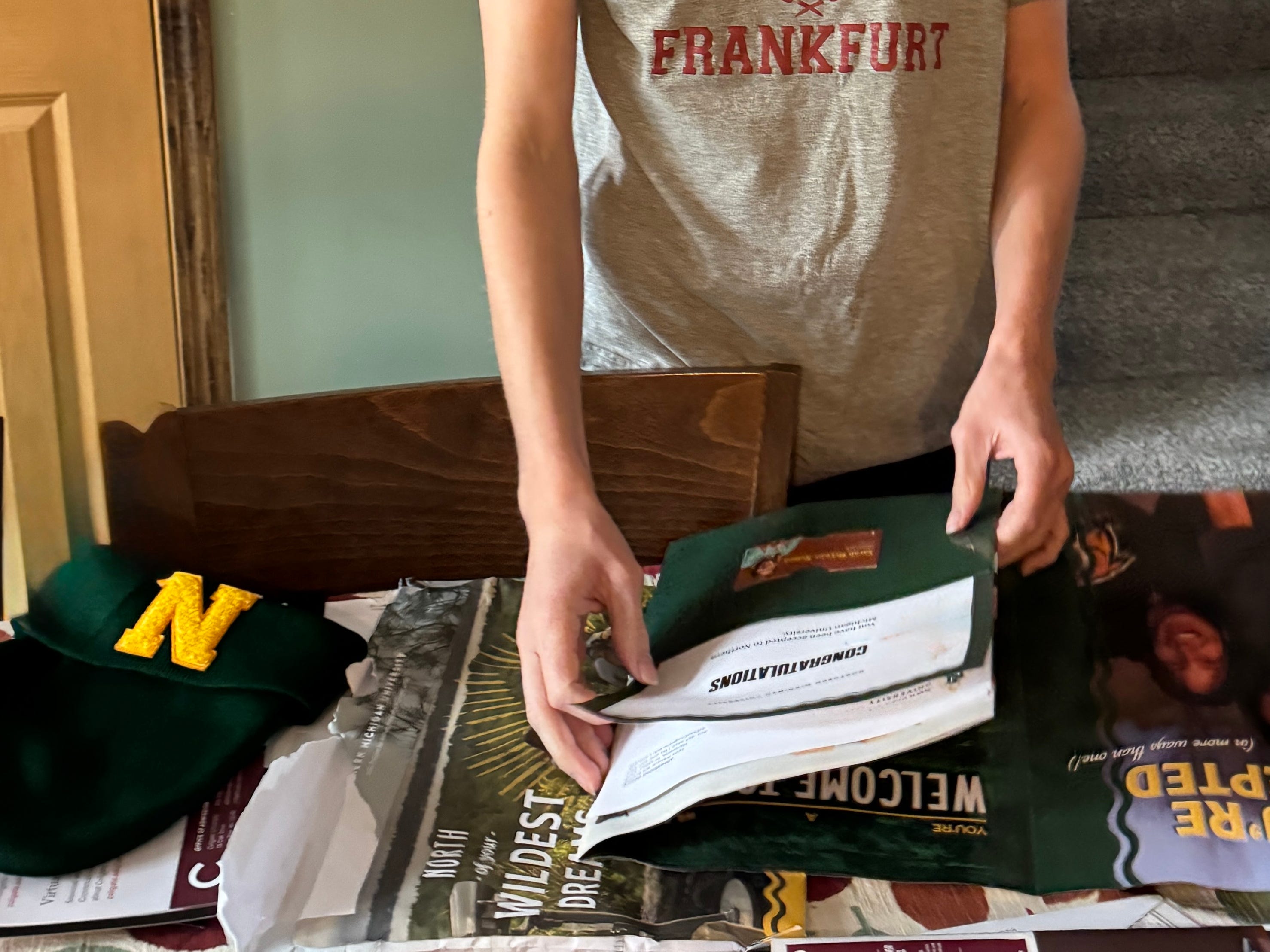 Person in a FRANKFURT shirt reviews college admissions materials beside a green cap with a yellow N.