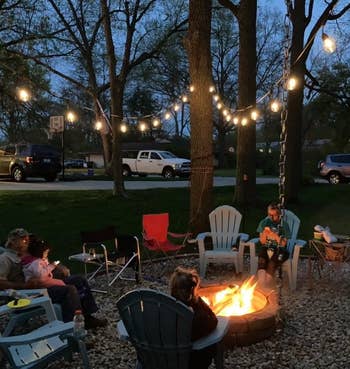 Family seated around a fire pit in a backyard at dusk, under string lights, with trees and parked vehicles in the background