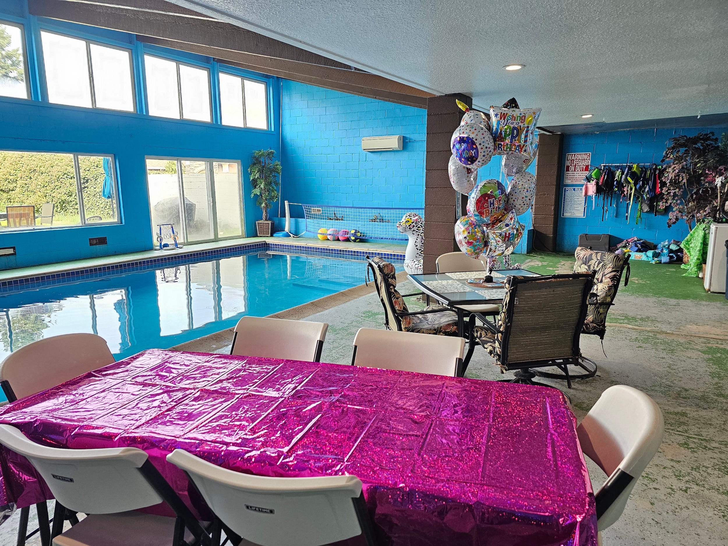 Indoor pool room decorated for a birthday party with balloons, folding chairs, and a pink tablecloth.