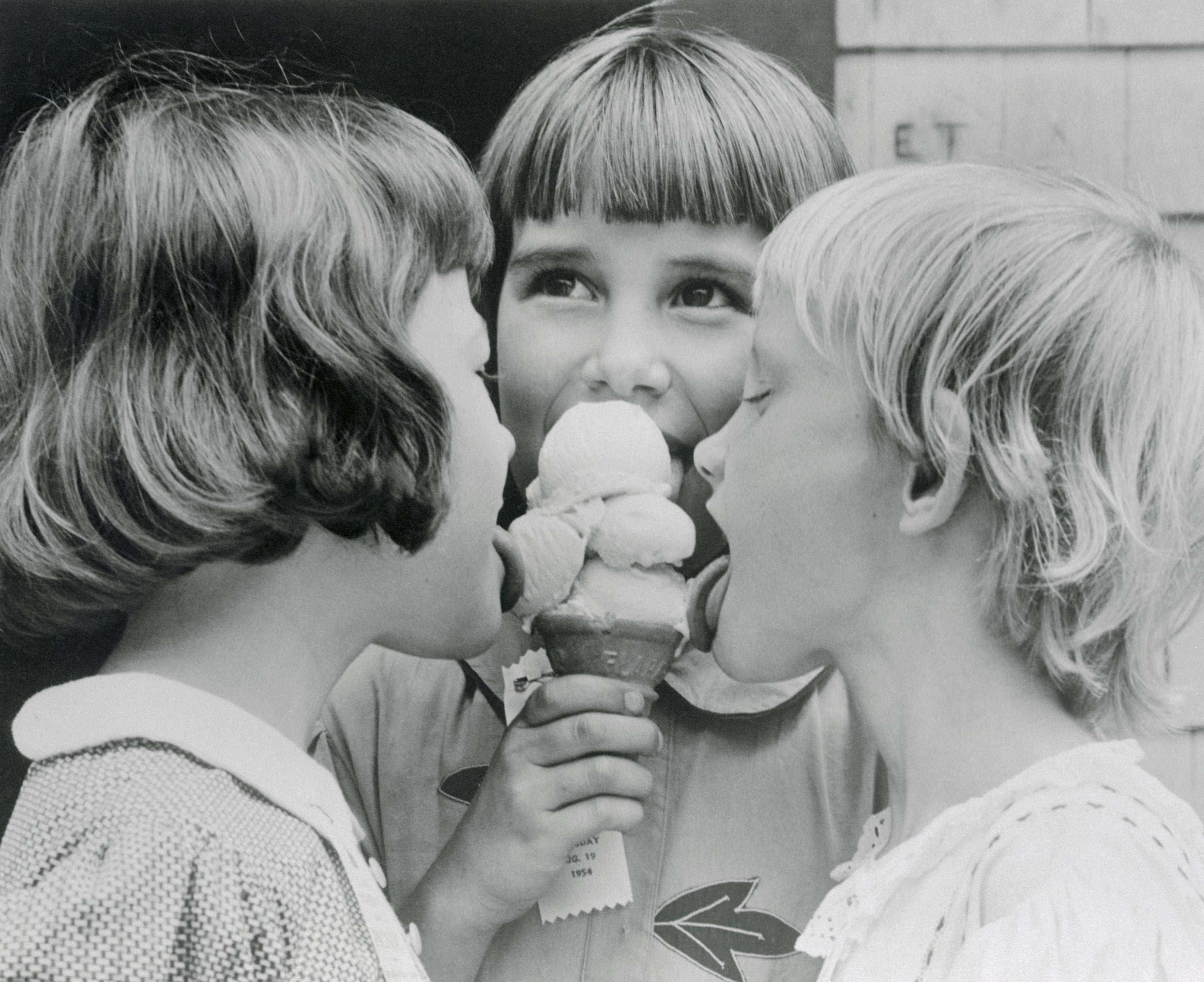 Three children sharing an ice cream cone, each taking a turn to lick it. They appear happy and playful
