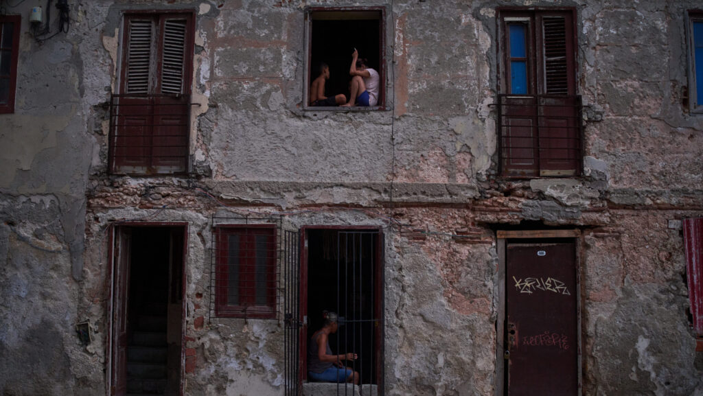 Two people sit in an upper window and another in a doorway of a crumbling, graffiti-marked building in Havana, Cuba