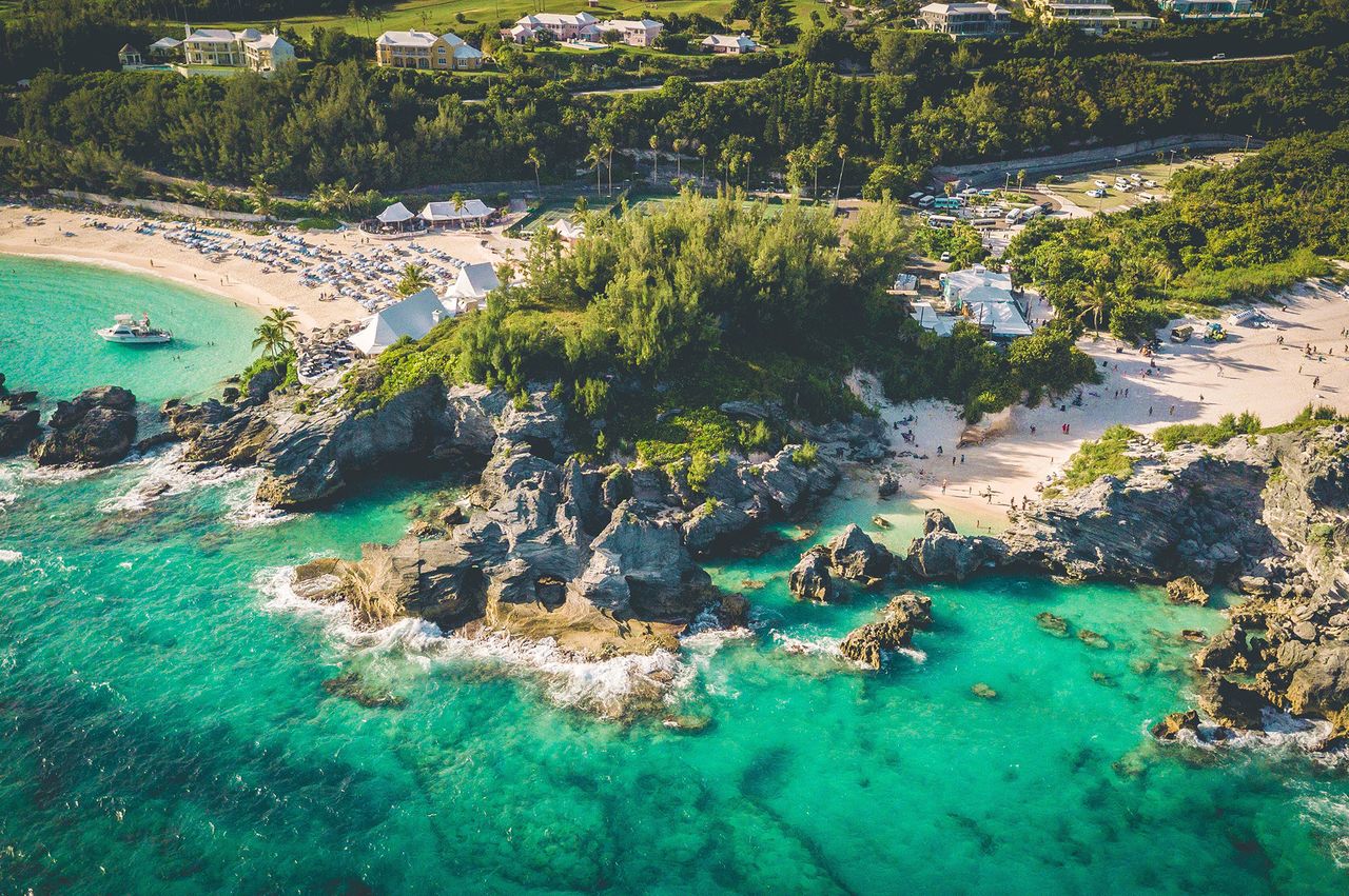 An aerial view of a scenic beach with surrounding greenery and rocky formations and small buildings visible near the shore