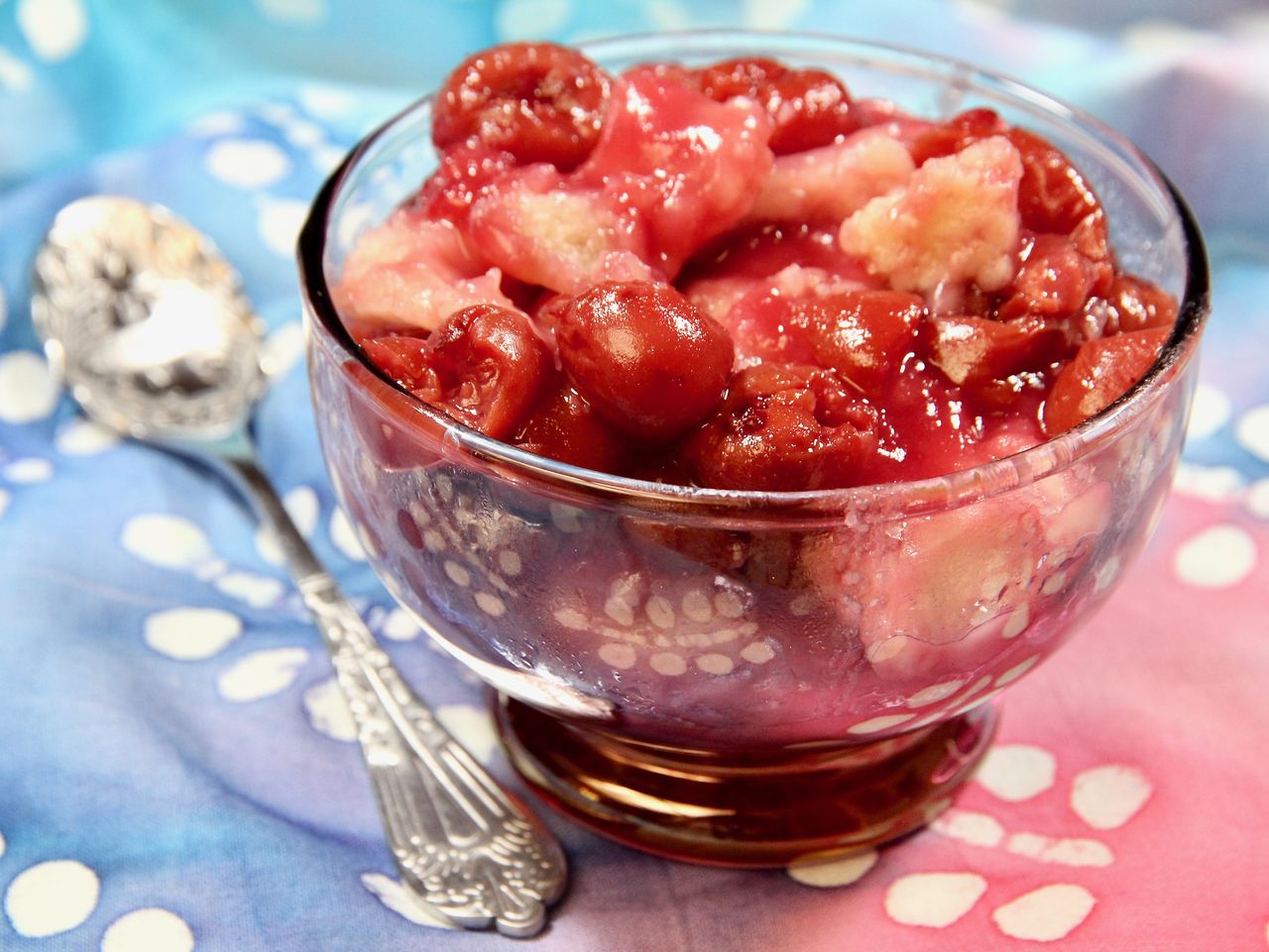 cherry cobbler in glass bowl with spoon on blue and red table cloth