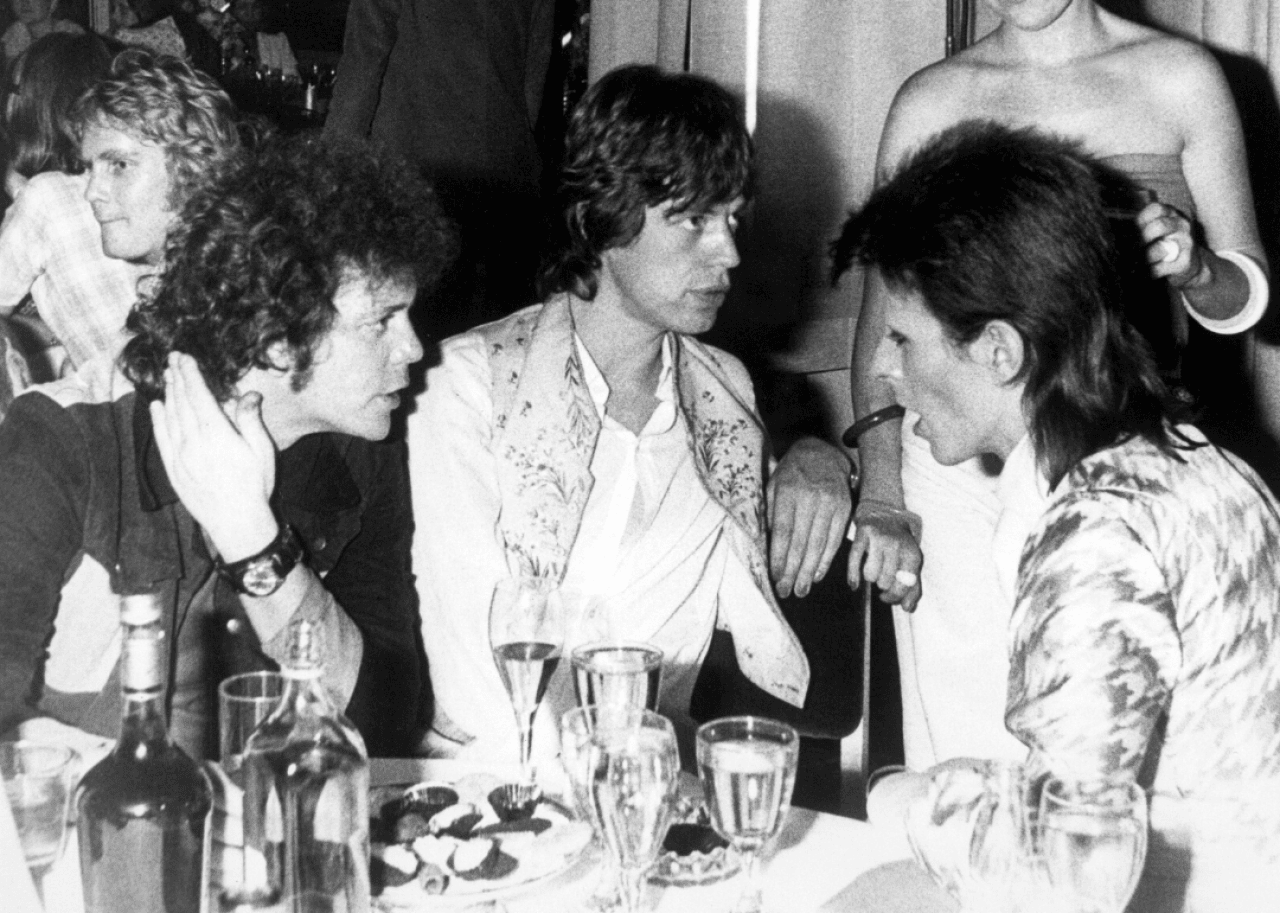 Lou Reed, Mick Jagger, and David Bowie sit together at a table in a restaurant.