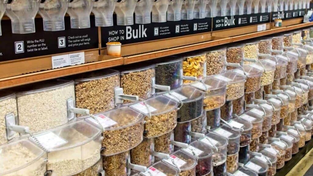 Bulk food dispensers in a store, filled with various grains, nuts, pasta, and dried goods, organized in clear containers with labels and instructions above.