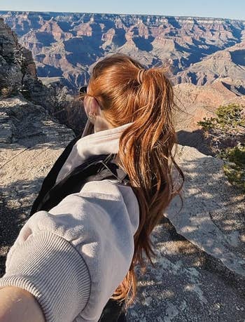 Person with long hair in ponytail taking a selfie on a rocky cliff with a vast canyon landscape in the background