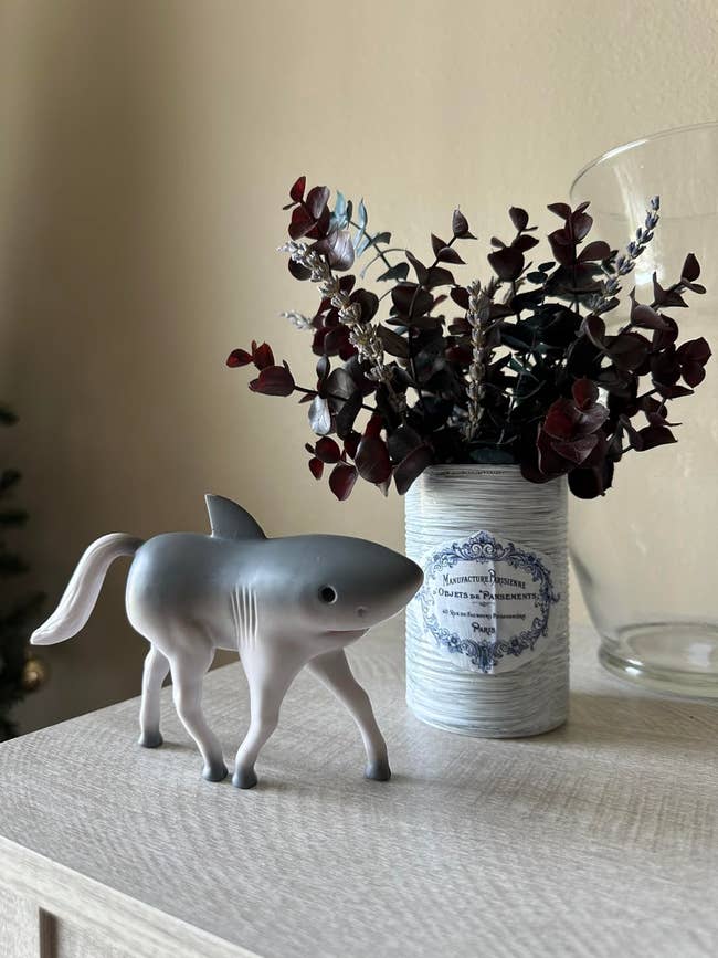 A whimsical figurine of a shark with legs stands on a table next to a decorative tin holding dark foliage