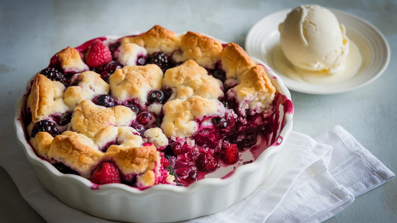 A berry cobbler in a white dish with one scoop missing and a big scoop of vanilla ice cream resting in a saucer in the background