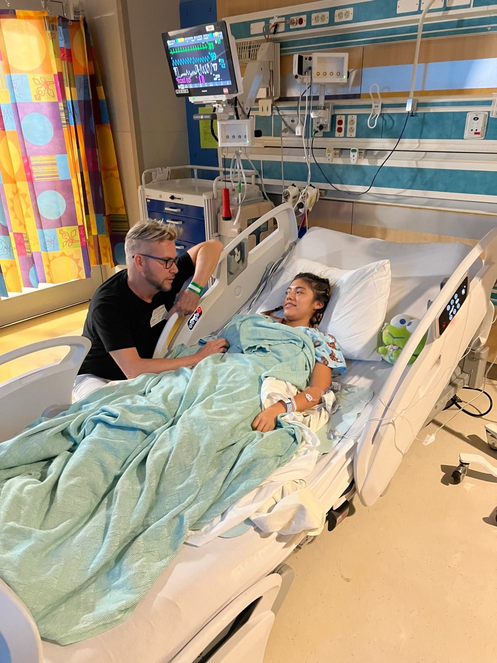 Armando Rubio sitting by Hannah Rubio's hospital bed, with a monitor displaying vital signs above.