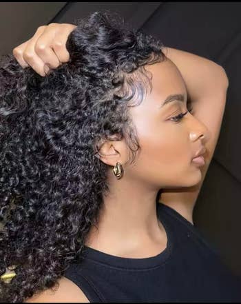 Person with curly hair in profile view, wearing a black top and hoop earrings, hand holding back hair
