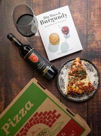 Pizza slice on a plate, open wine bottle, glass of red wine, pizza box, and wine-and-burger pairing book on a wooden table