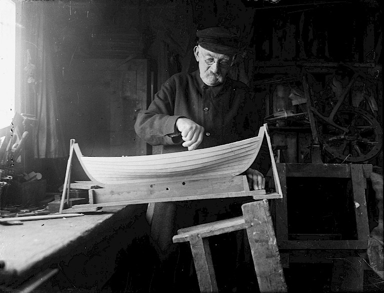 An elderly man in a flat cap and glasses works on the curved wooden hull of a model boat in his workshop.
