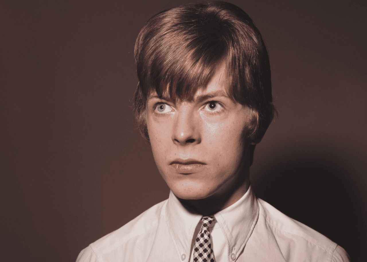 A portrait of a teenage Bowie in a white button up shirt and tie.