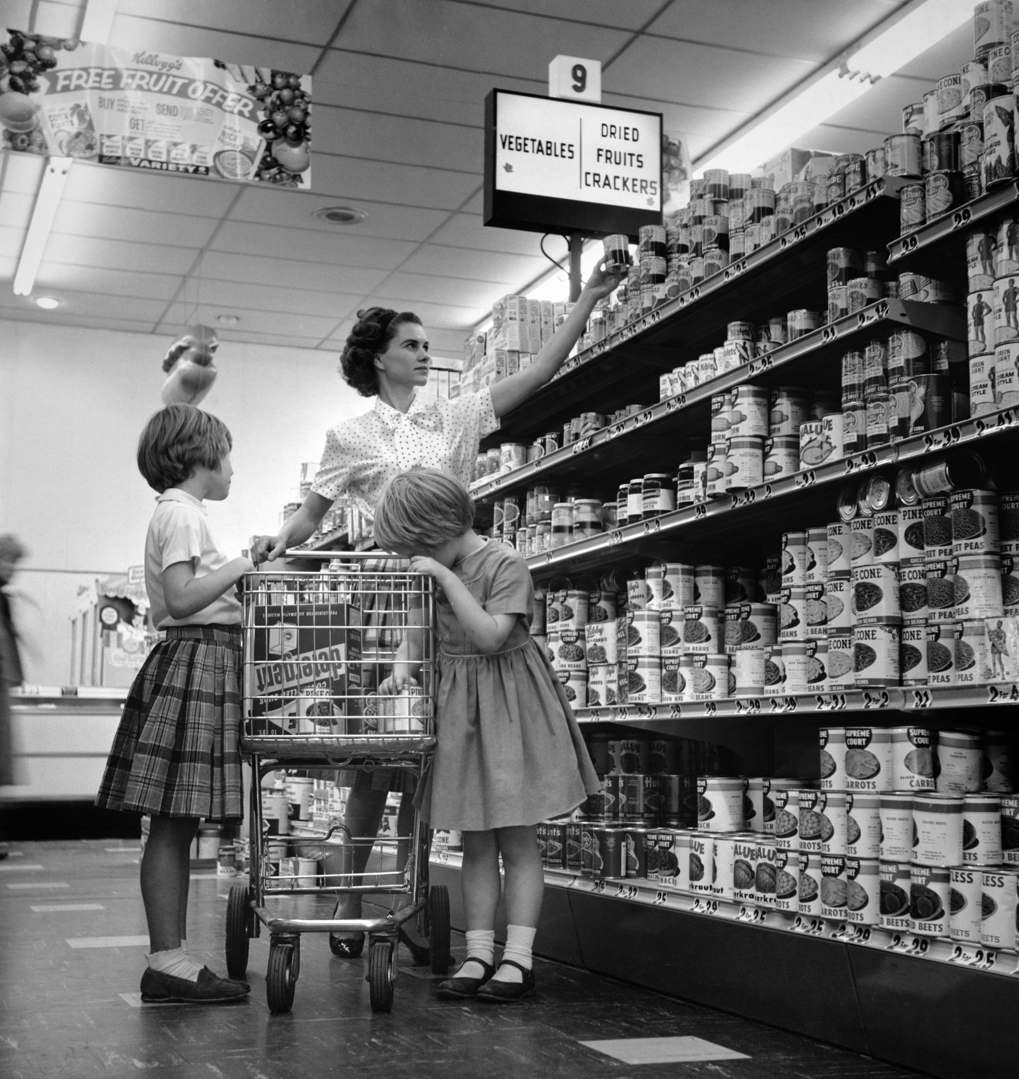 Woman and two girls shopping in a grocery store aisle, selecting canned goods from shelves