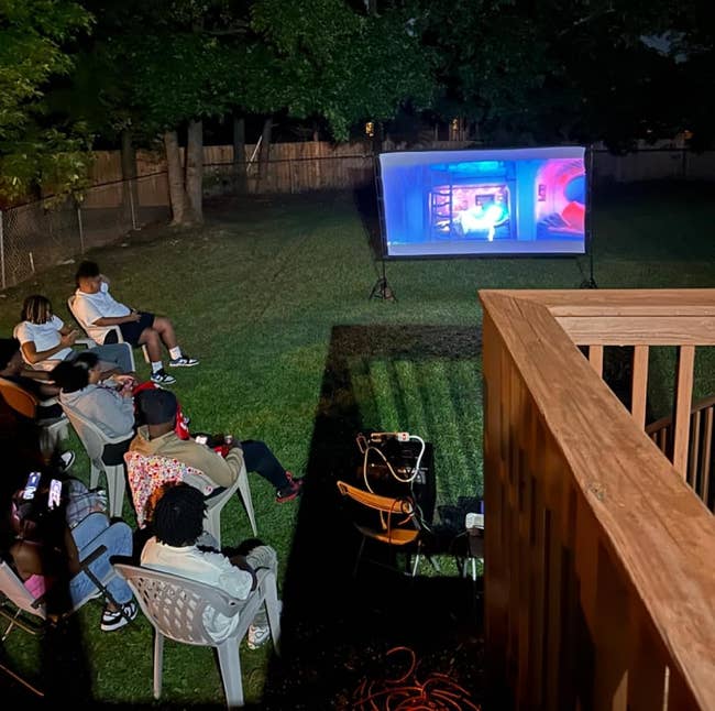 Outdoor movie night setup with several people seated in chairs watching a projected film in a backyard. Trees and a fence are visible