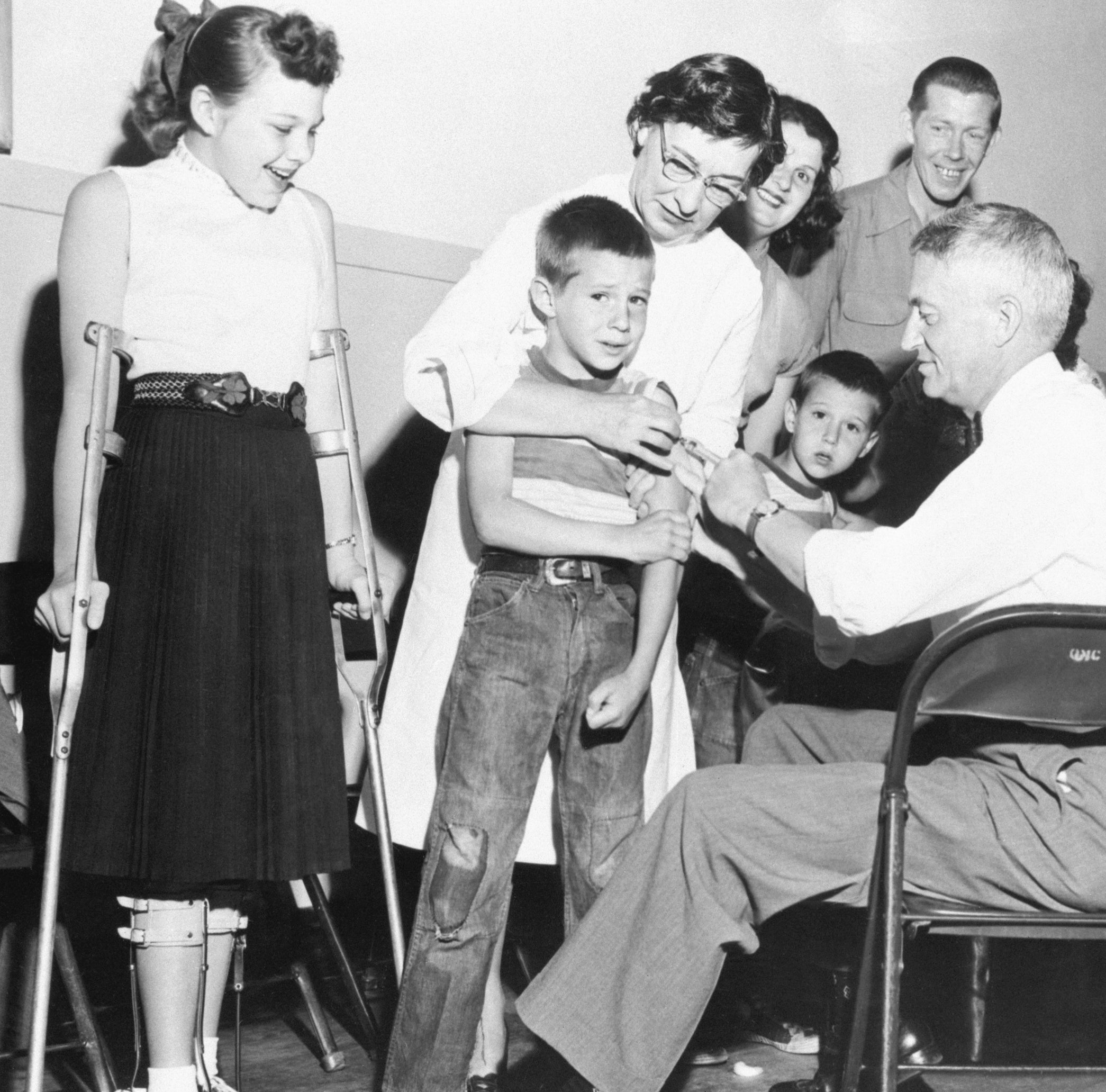 A young boy receives a shot while others, including a girl with crutches and medical staff, look on in an early vaccination clinic scene