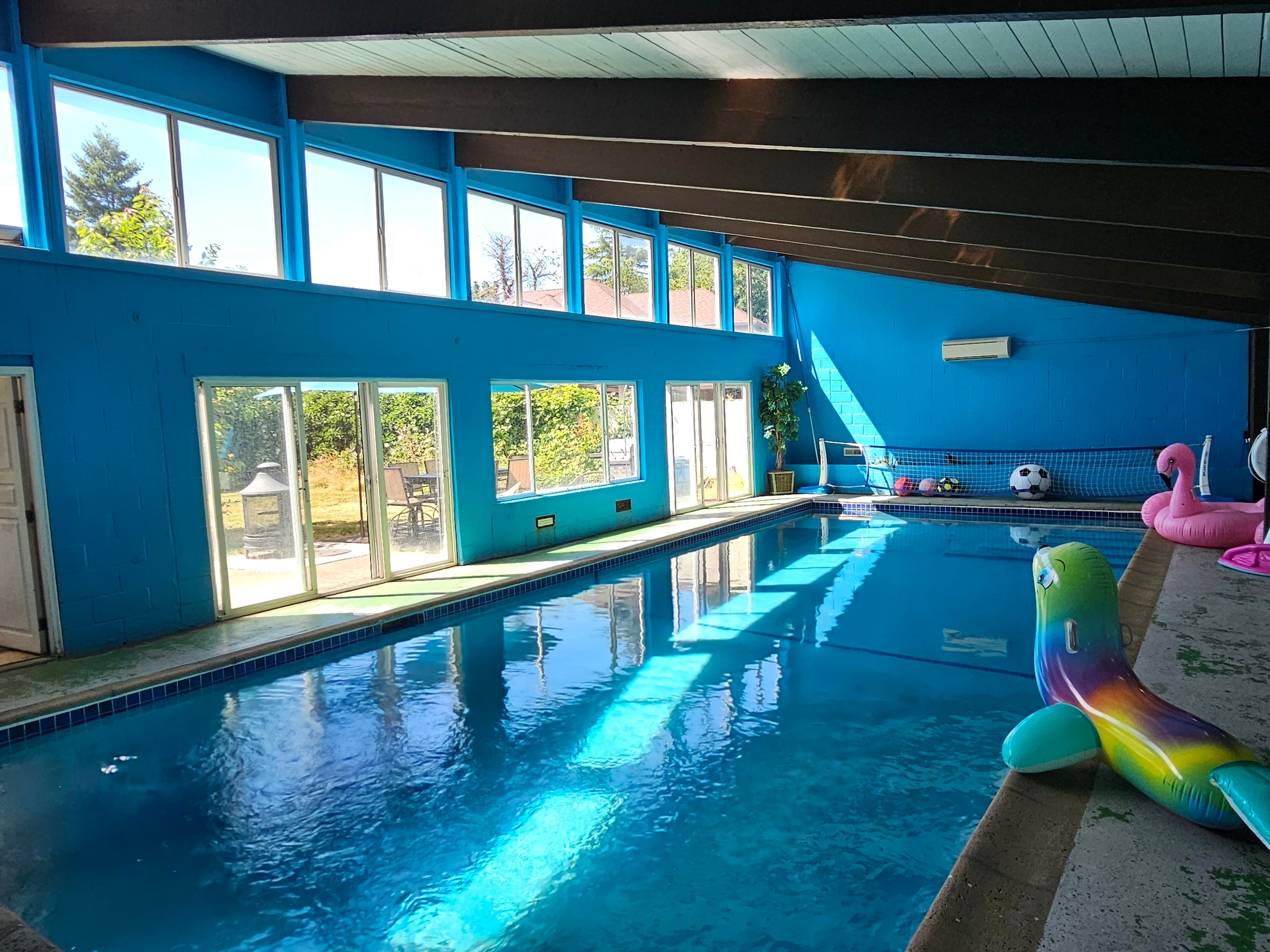 Indoor swimming pool with bright blue walls, high windows, sliding doors, and colorful inflatable pool floats.