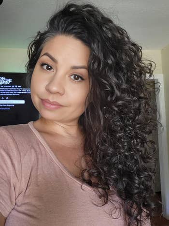 Person with wavy hair and neutral expression wearing a casual top. TV screen in the background displays movie options