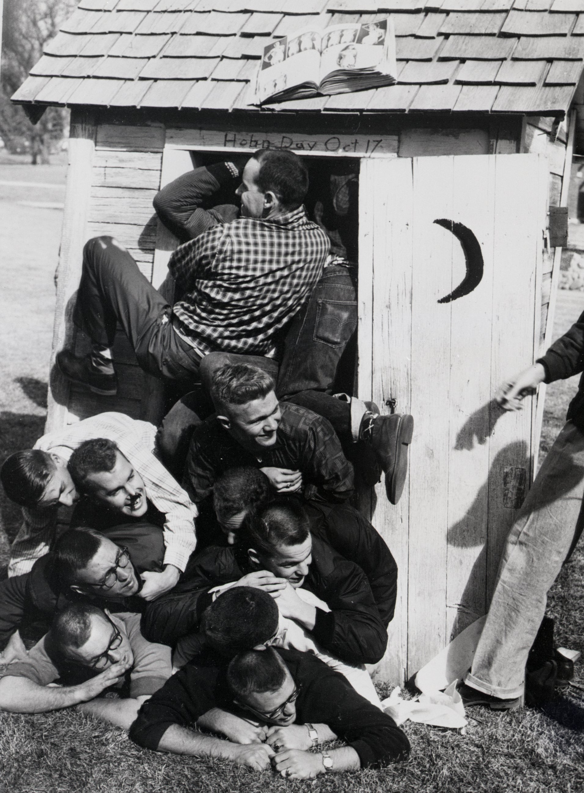Seven young men humorously cram into an outhouse on a college campus on Hobby Day, with a small wooden structure and a crescent moon on the door