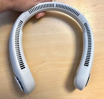 Hand holding a wearable neck fan with vent openings, suitable for hands-free cooling and featured in a shopping article