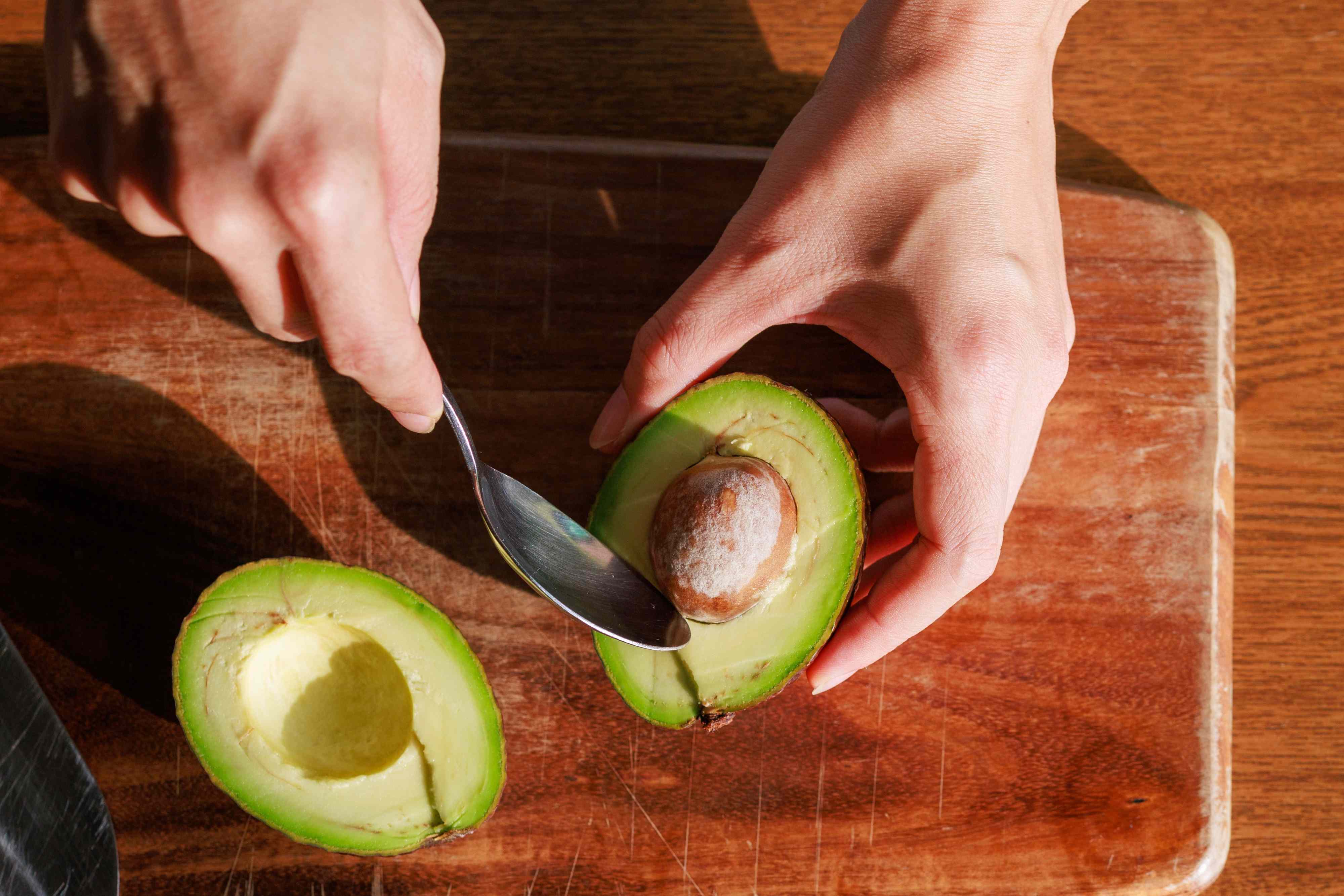 Person scooping an avocado half with a spoon on a wooden surface
