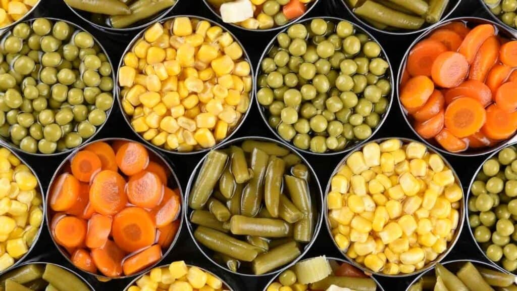 Open cans filled with canned vegetables, including corn, peas, carrots, and green beans, arranged closely together and viewed from above.