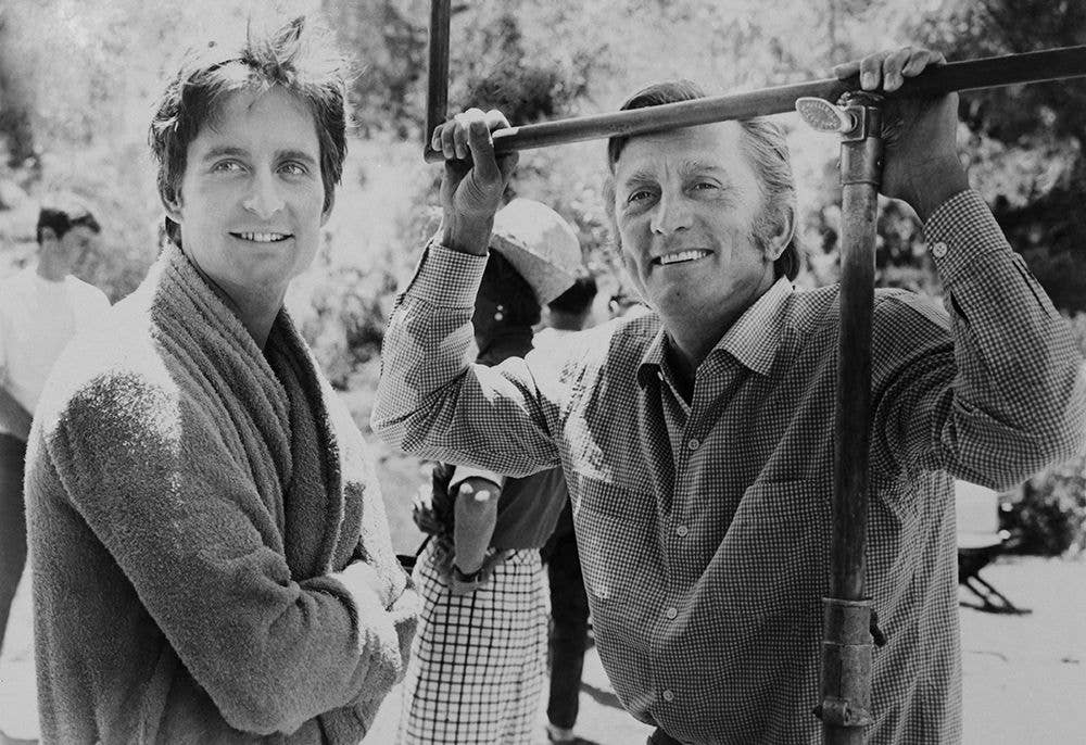 Michael Douglas in a robe and Kirk Douglas in a checkered shirt, both smiling and holding a bar.