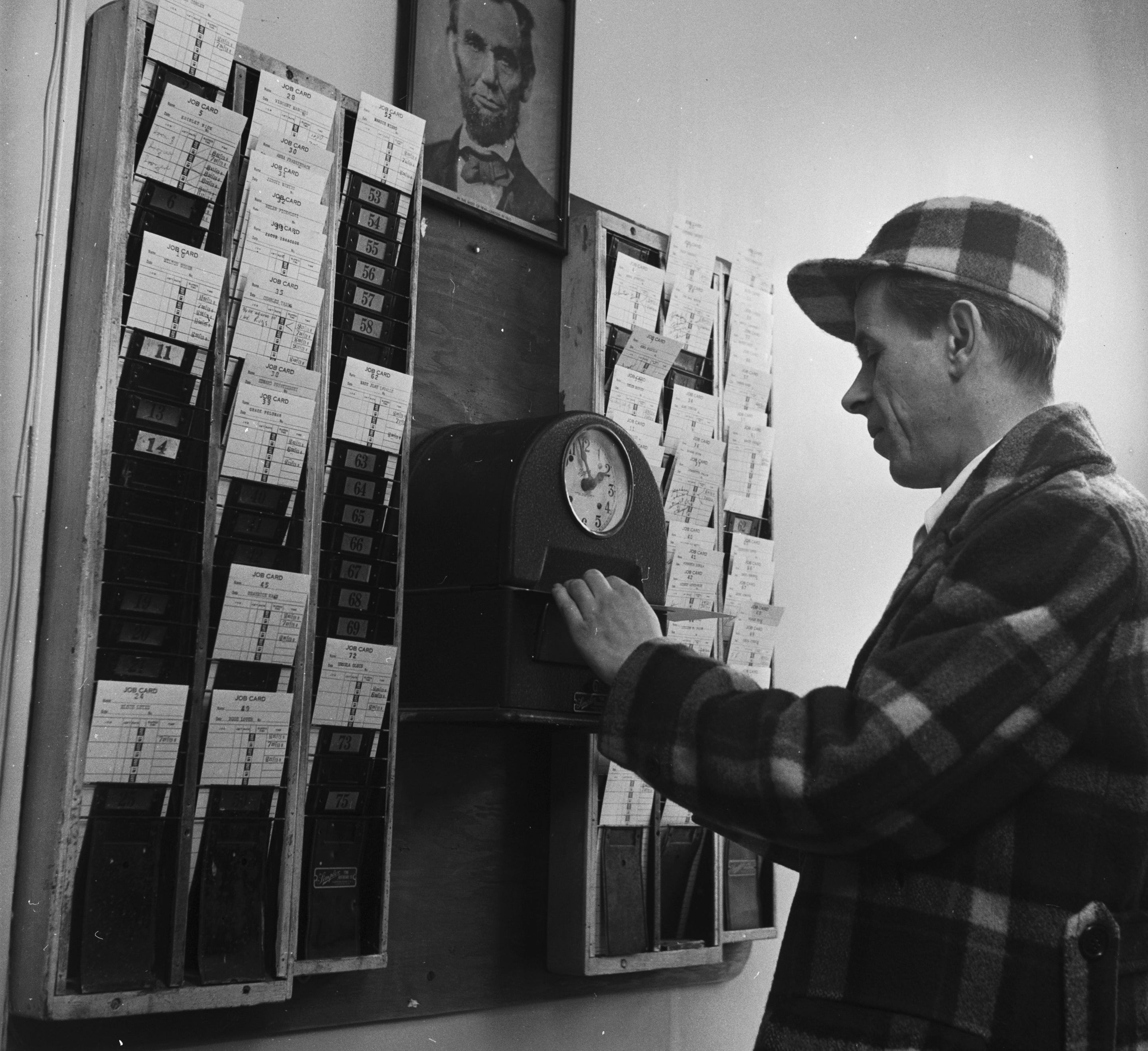 A man in a plaid coat punches a timecard at a clock beneath a portrait of Abraham Lincoln in an office setting