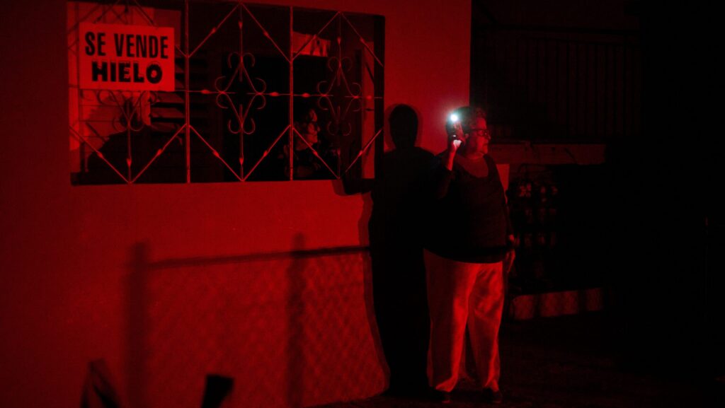 Two people stand outside a home at night, lit in red, next to a barred window with a sign reading