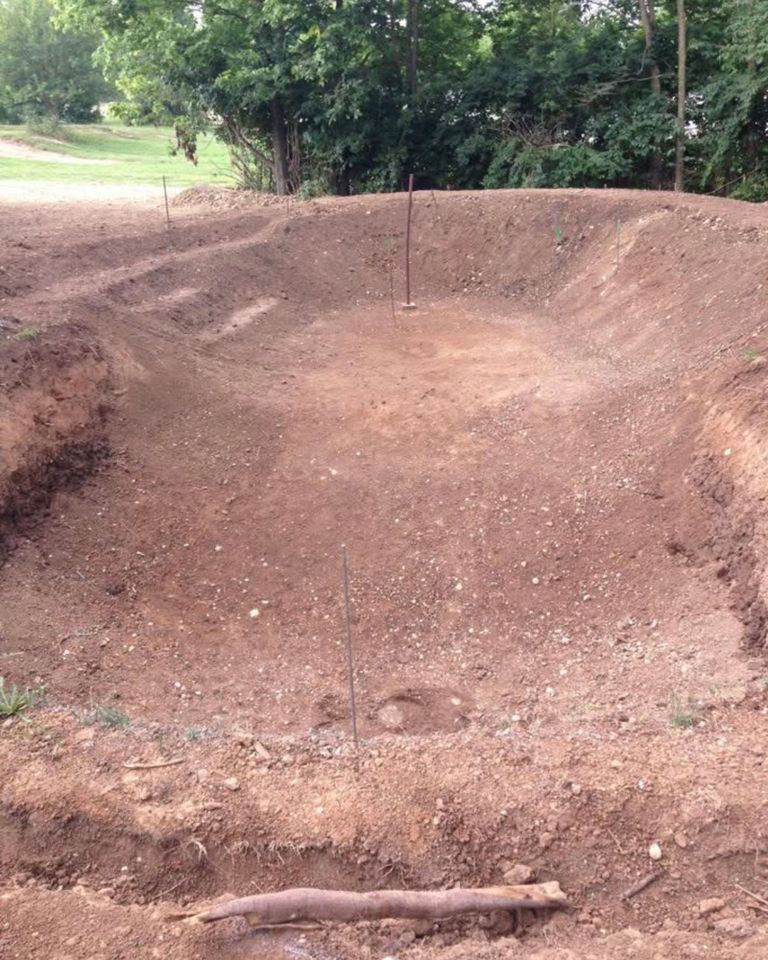 Excavated ground prepared for a pond or pool surrounded by soil and greenery in the background