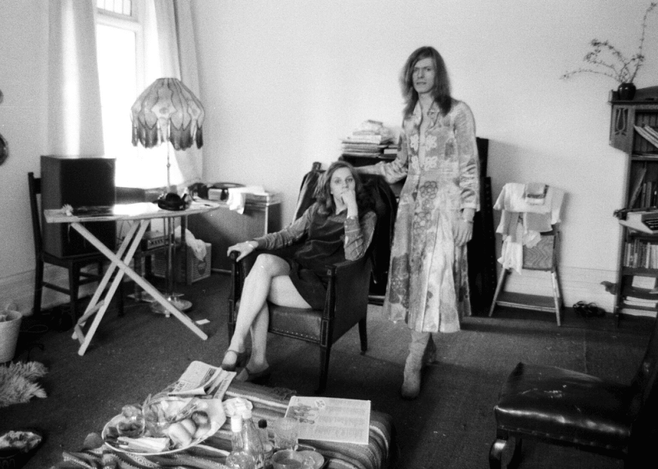 Angela Barnett sits in an arm chair with David Bowie in a dress standing beside her.