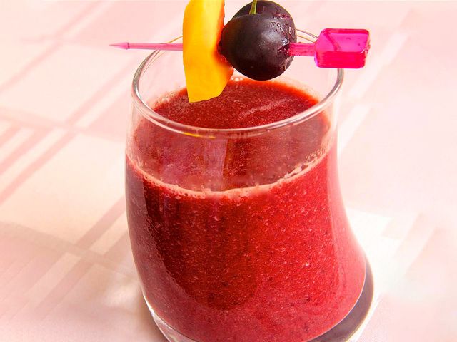 close up view of a Mango Cherry Smoothie in a glass, garnished with a cherry and a piece of mango on a pink toothpick that's on top of the glass